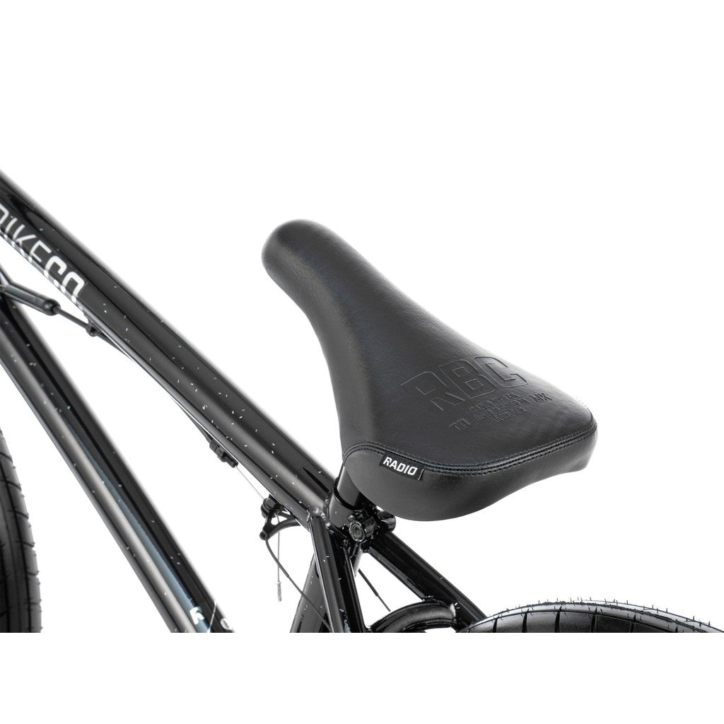 Close-up of a black bicycle seat with the brand "Radio" visible on the side. The frame and rear tire of the Radio Astron FS 20 Inch Bike, featuring a full chromoly frame and Radio Pro Hubs, are partially visible.