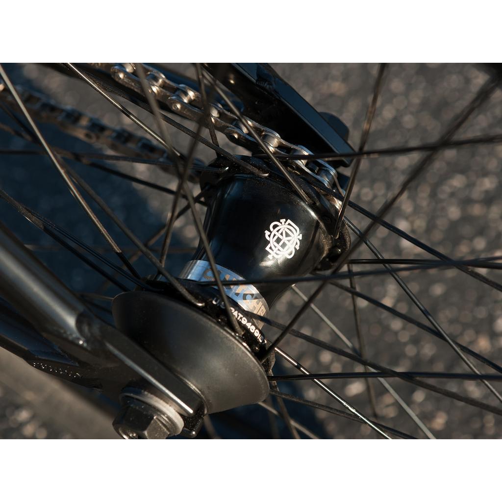 Close-up of a Sunday Sound Wave Special 20 Inch Bike (2023) rear wheel hub, showcasing Odyssey parts, with spokes, chain, and logo details set against a blurred asphalt background.