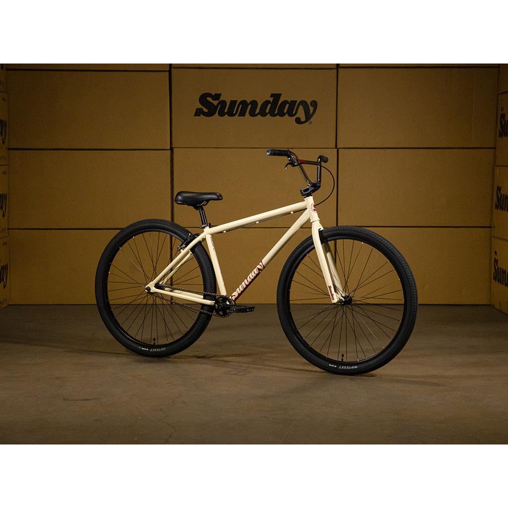 The Sunday High C 29 Inch Bike—a cream-colored BMX-style bicycle with black tires—stands indoors before stacked cardboard boxes marked "Sunday." This 29" cruiser boasts a 100% chromoly frame for both superior strength and timeless style.