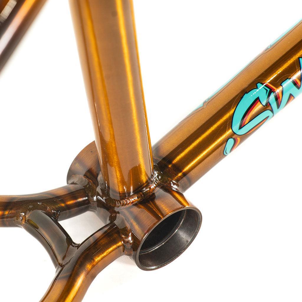 Close-up of a metallic bronze bike frame junction with clean welds, featuring the Colony 2024 Sweet Tooth Frame logo in blue and white on its chromoly construction.