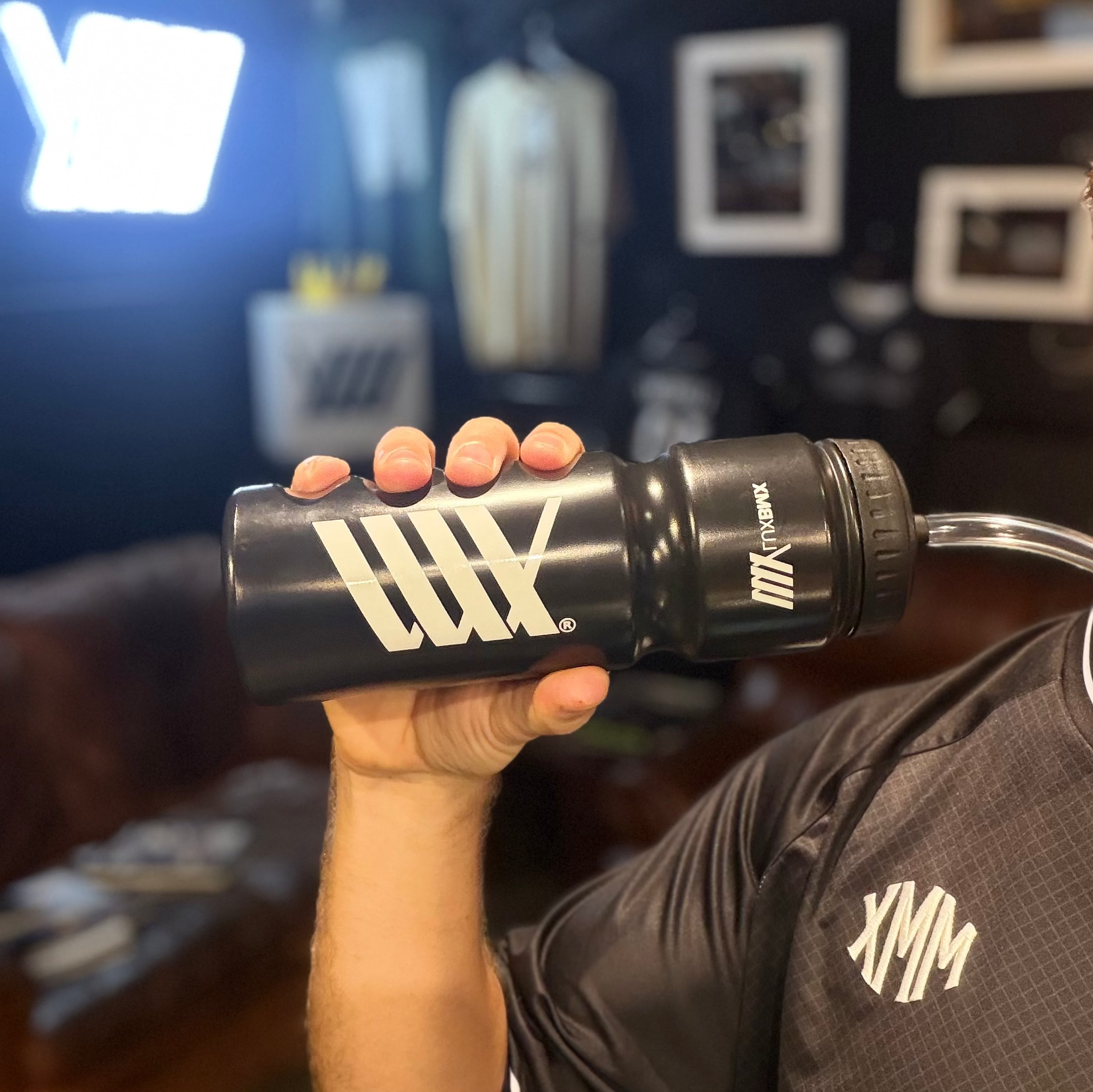 A person in a black XMM shirt holds a 750ml LUXBMX Race Straw Water Bottle made from BPA-free plastic, preparing to drink indoors with framed items visible on the wall.