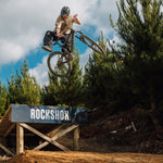 A person wearing a helmet and the Buzzed Riders Split Tee catches air on a mountain bike over a wooden ramp branded with "RockShox," surrounded by trees and partly cloudy skies—an action-sport moment that's sure to spark conversation.