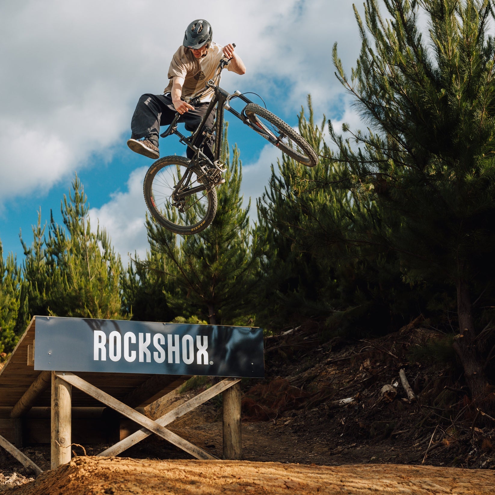 A person wearing a helmet and the Buzzed Riders Split Tee catches air on a mountain bike over a wooden ramp branded with "RockShox," surrounded by trees and partly cloudy skies—an action-sport moment that's sure to spark conversation.