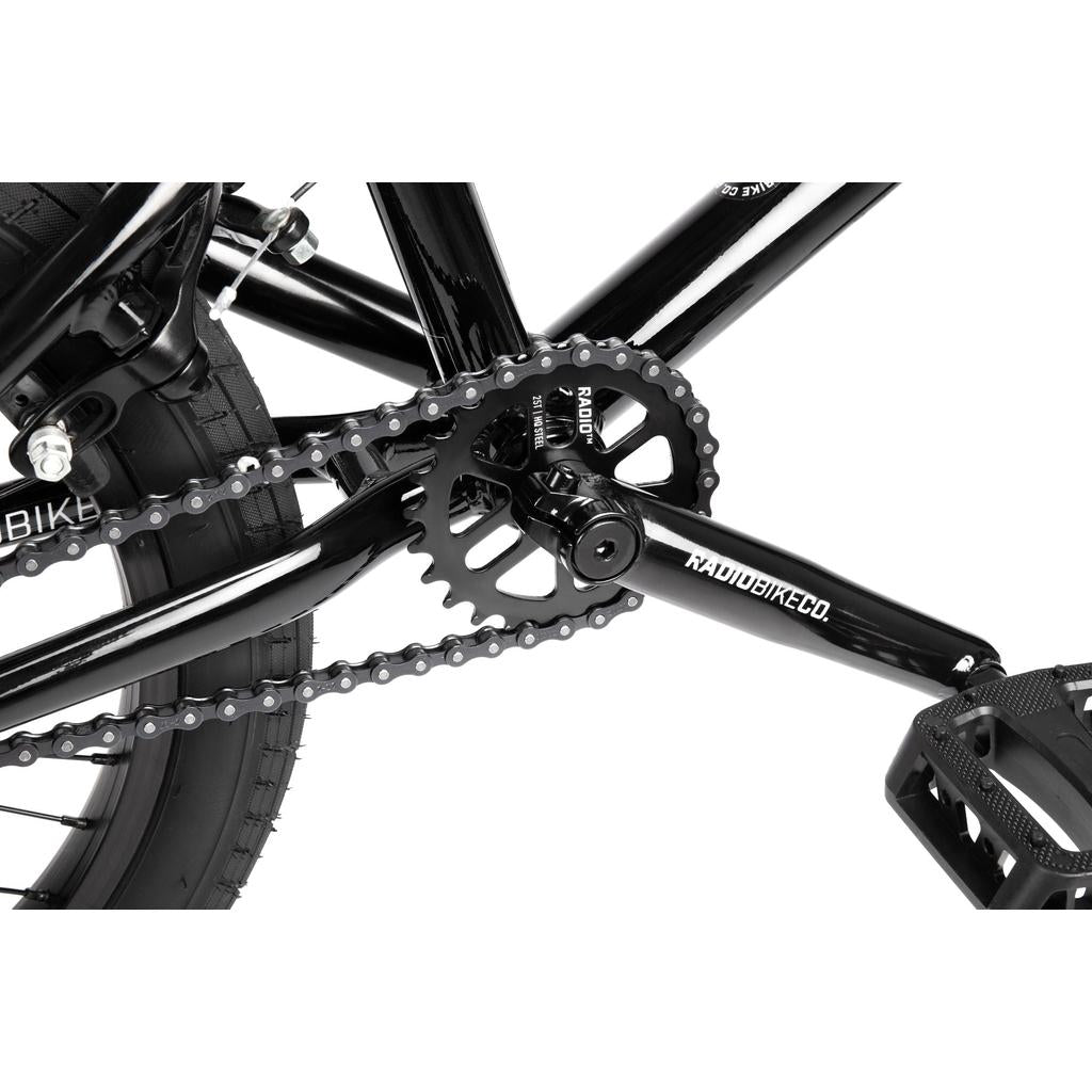 Close-up of the drivetrain on a black Radio Dice 18 Inch Bike, showing the chain, crank arm, chainring, pedal, part of the rear wheel, and Radio Rookie Three Piece Cranks.