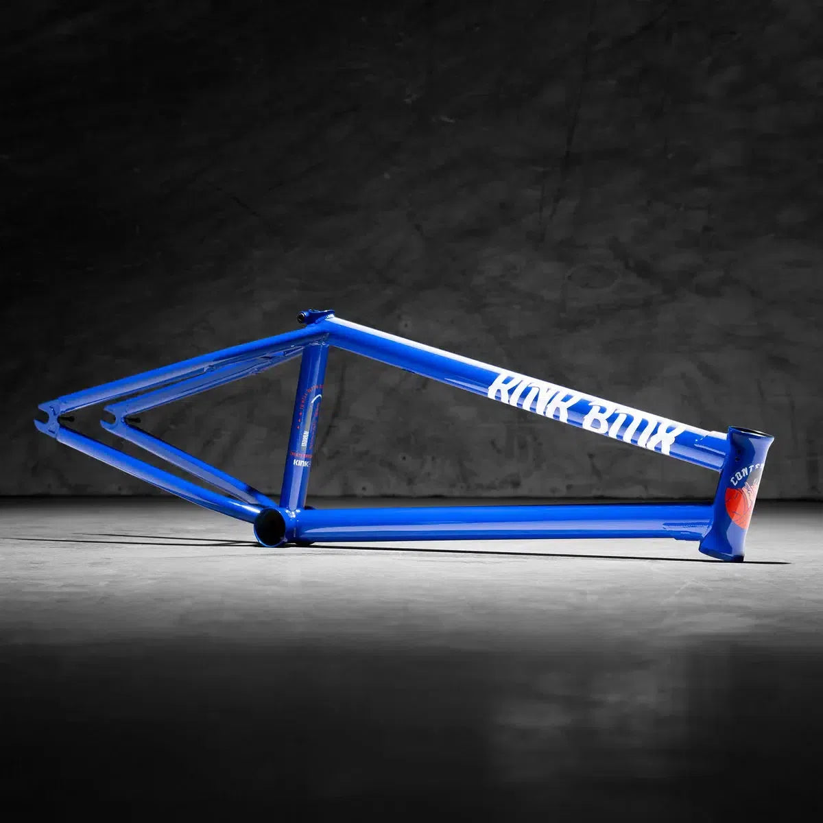 The Dan Coller signature Kink Contender 2 blue BMX bike frame, labeled "RIDE BLUE," rests on a gray concrete floor against a dark backdrop, epitomizing durability and style.