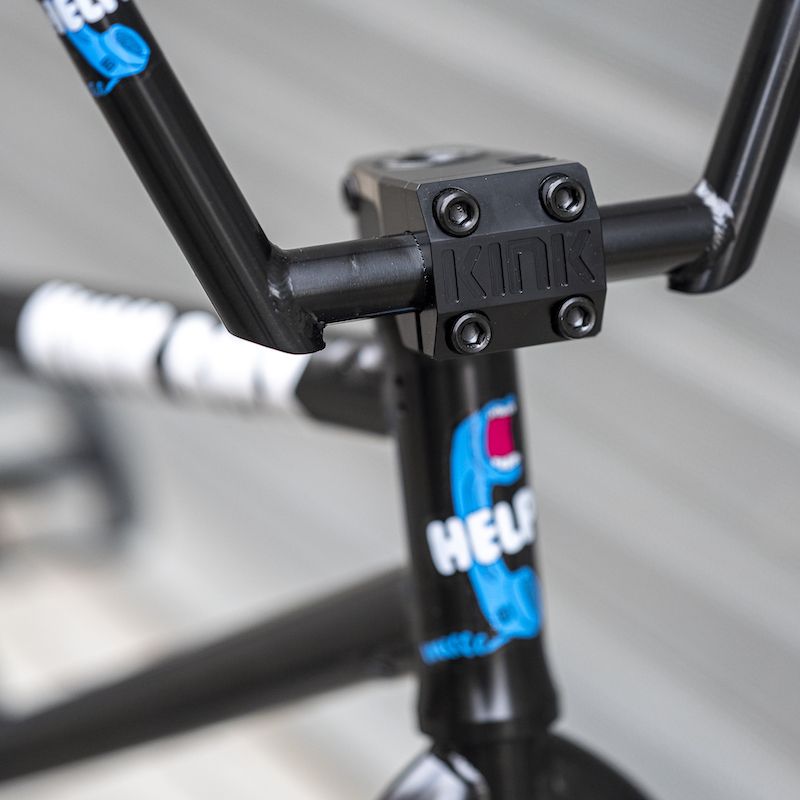 A close-up of a black BMX bike handlebar highlights the CNC machined Kink Track Front Load Stem, featuring vibrant decals and an engraved KINK logo. The detailed craftsmanship emphasizes "Help," adding character and style to the ride.