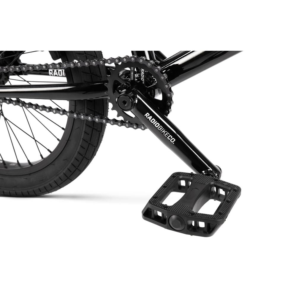 Close-up of a black BMX pedal, Radio Rookie Three Piece Cranks, chain, and Salt Tracer tyres on the rear wheel of the Radio Dice 18 Inch Bike against a white background.