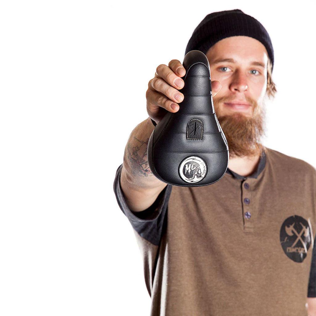 A person wearing a beanie and a brown logo shirt holds up the Demolition Matt Cordova Seat (V1) towards the camera, striking a pose reminiscent of Matt Cordova's iconic style.