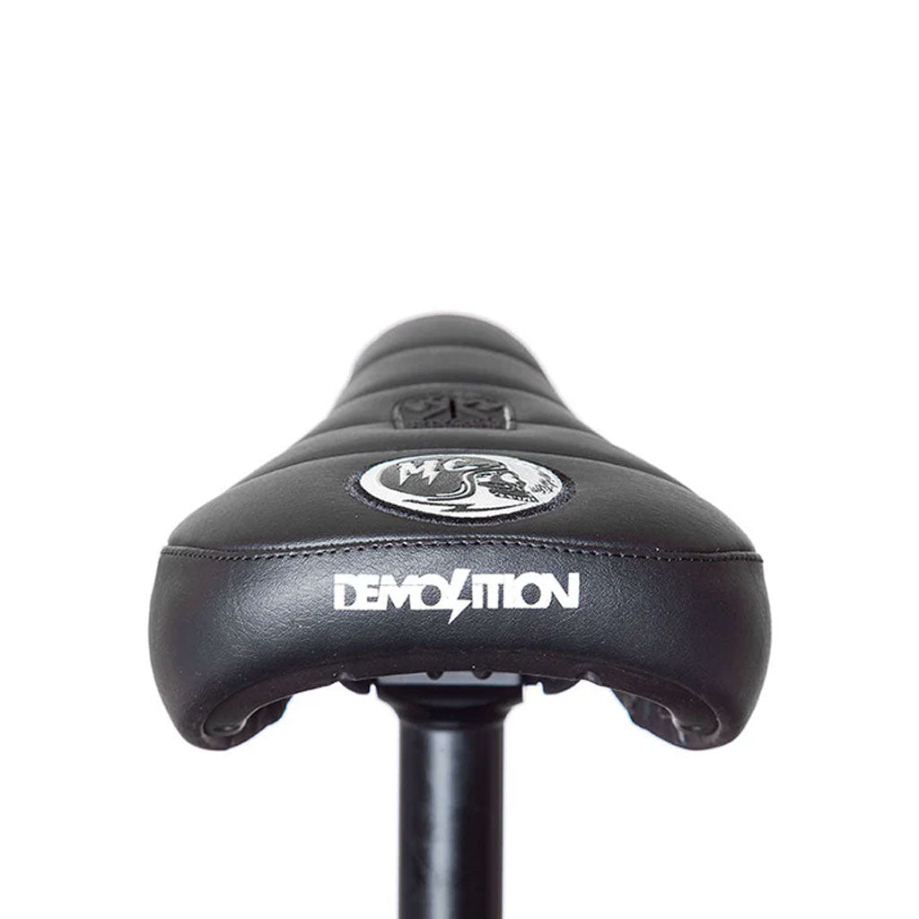 The Demolition Matt Cordova Seat (V1) is a black BMX seat with a white "DEMOLITION" logo on the back, showcasing the Pivotal Fat Cap design. It’s attached to a seat post against a plain background, highlighting Matt Cordova's impact on its style and functionality.