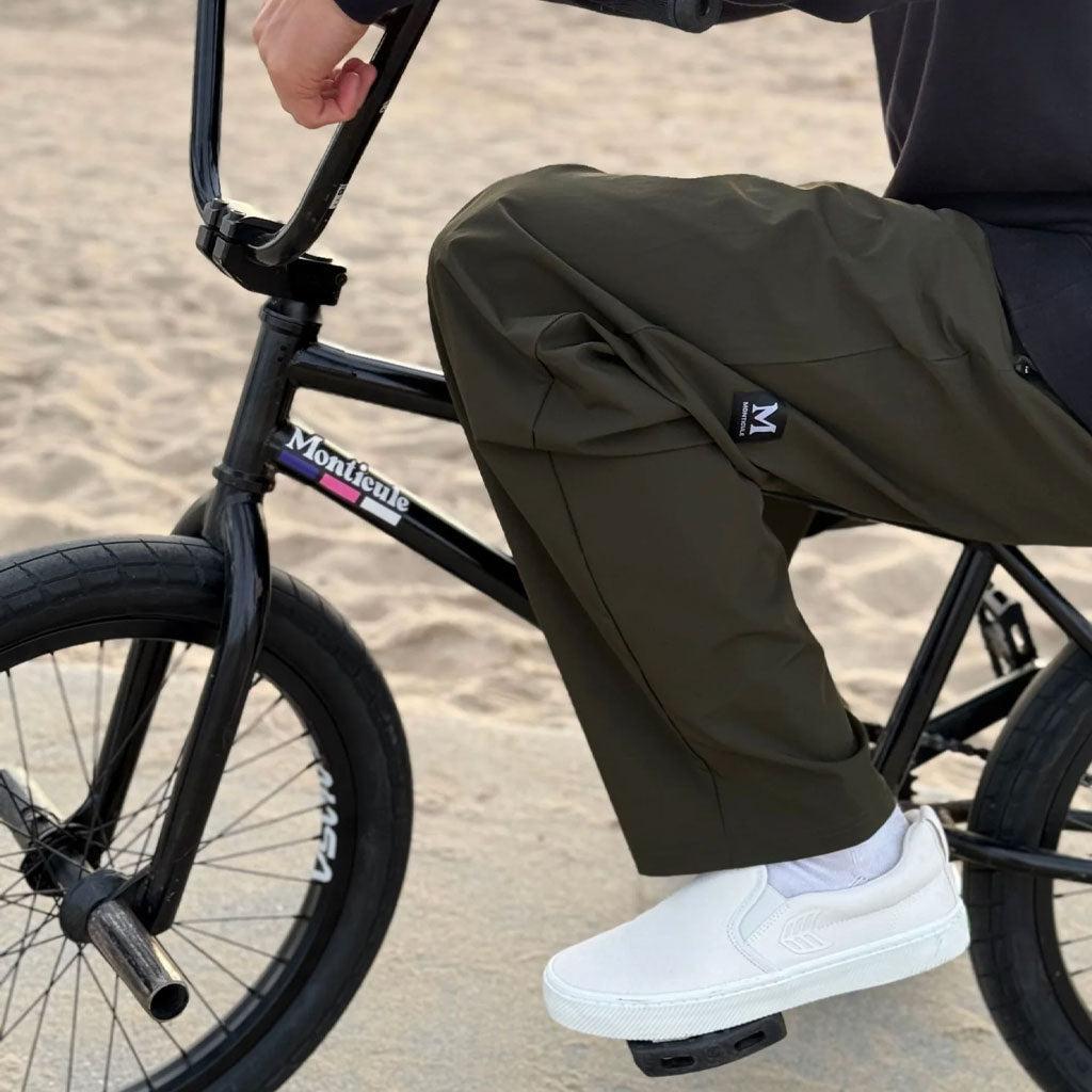 Dressed in Monticule Ghosts Pants and white slip-on shoes, the person sits on a black BMX bike on sandy ground.