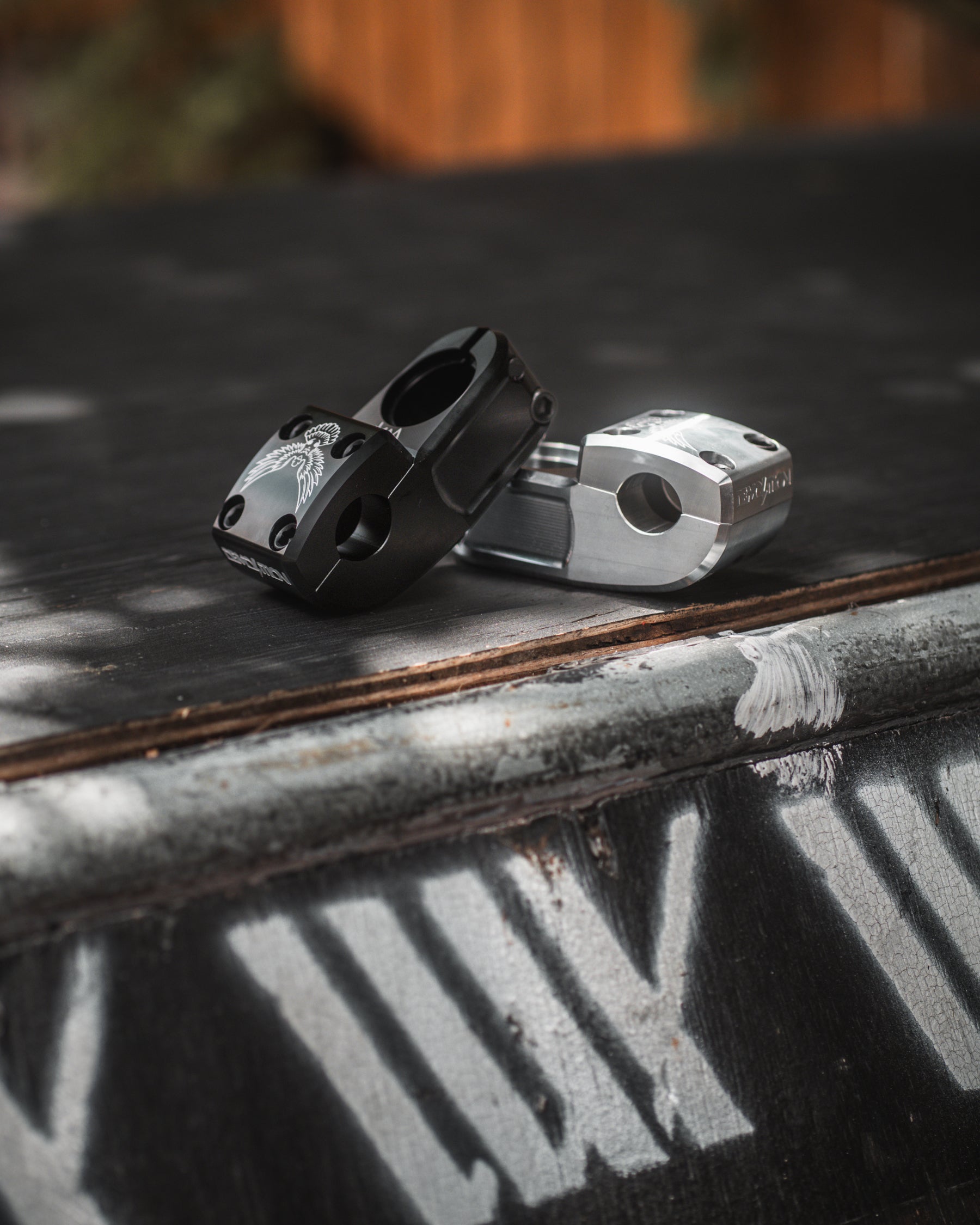 Two metal BMX bike stems, including the Demolition Josh Dove Stem, are displayed on a rough surface. One is black, and the other is silver, with stencil-like text in the background enhancing their rugged appeal.