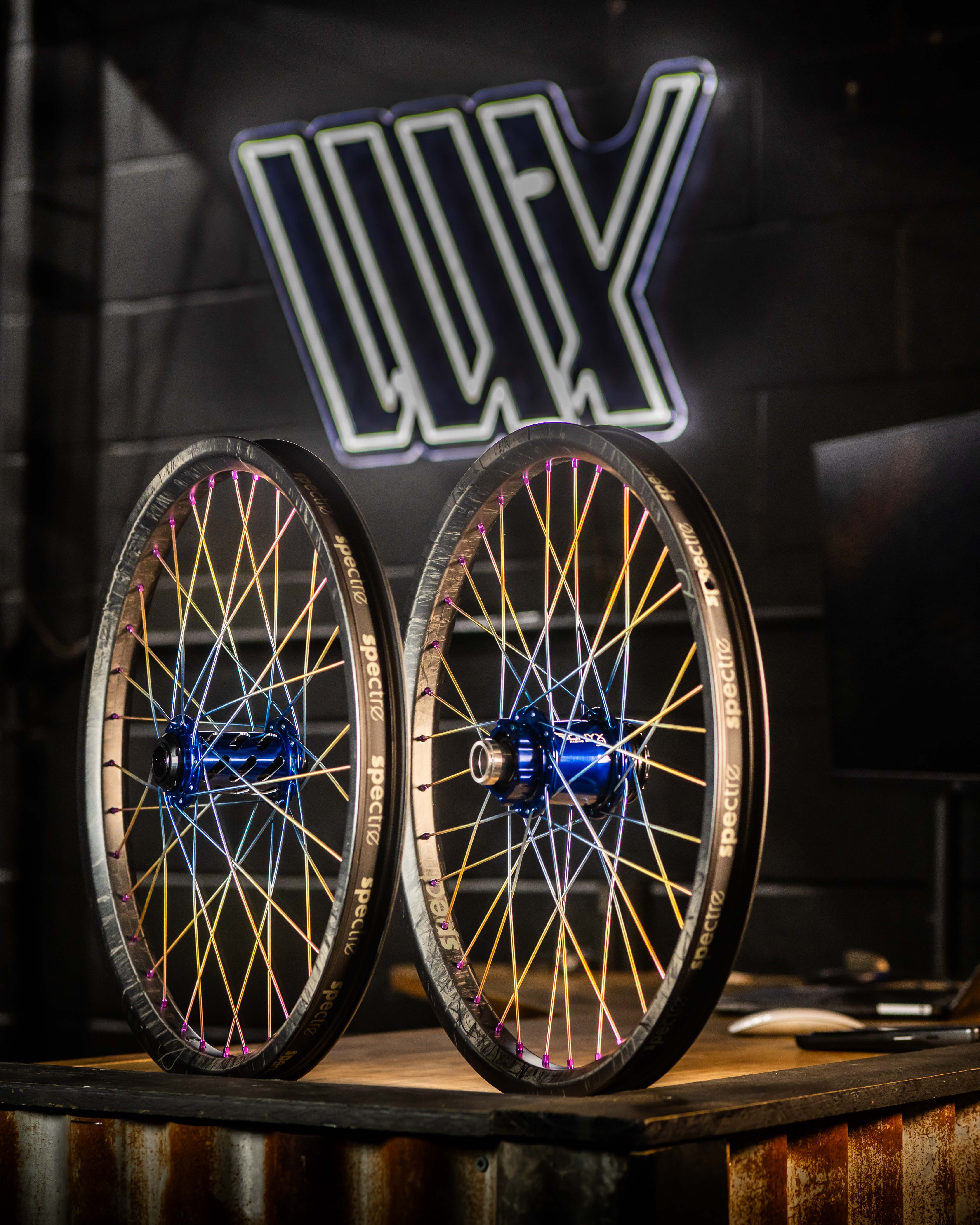 Two Onyx X Spectre Carbon Marble Sunset 20 Inch Wheels with colorful titanium spokes and blue hubs are upright on a wooden table against a dark wall featuring a large, illuminated logo.