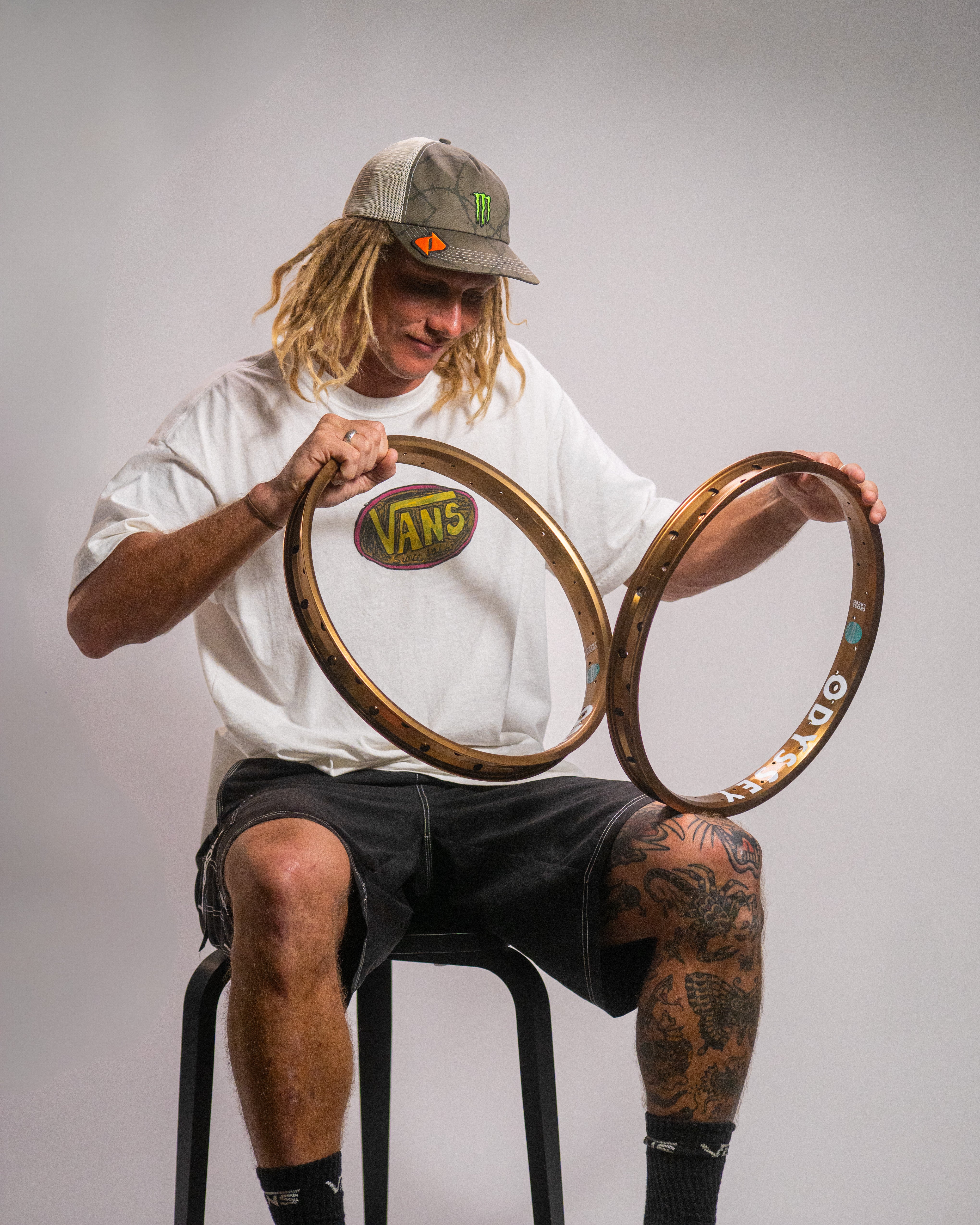 A person with long dreadlocks in a Vans t-shirt and cap sits on a stool, holding two Odyssey Big Boyd Rims—HARD ANODIZED BLACK with extra-wide cross-sections—set against a plain backdrop.