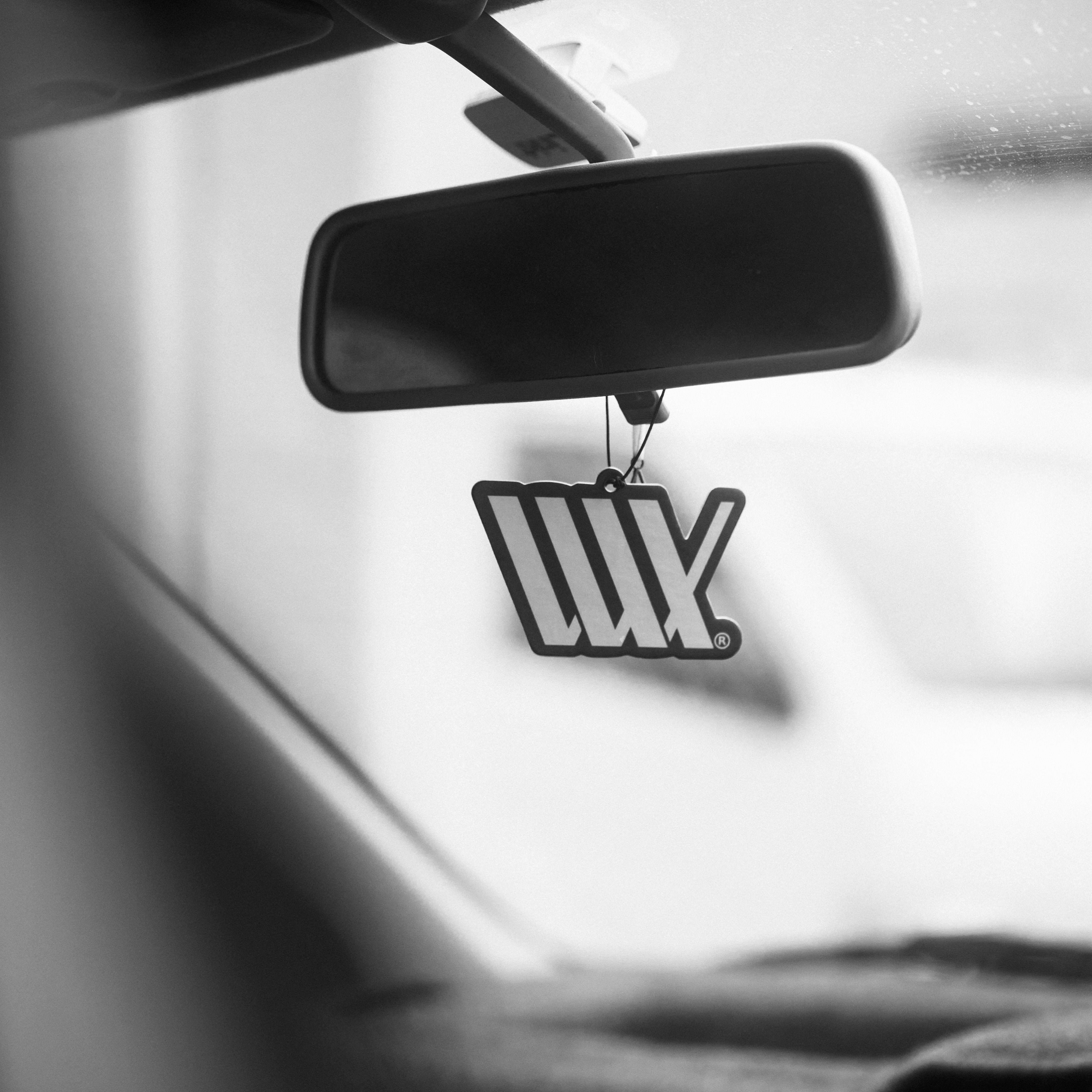 A black and white photo of a car's rearview mirror with a LUXBMX Air Freshener - Josh Dove hanging from it, capturing the essence of freshness.