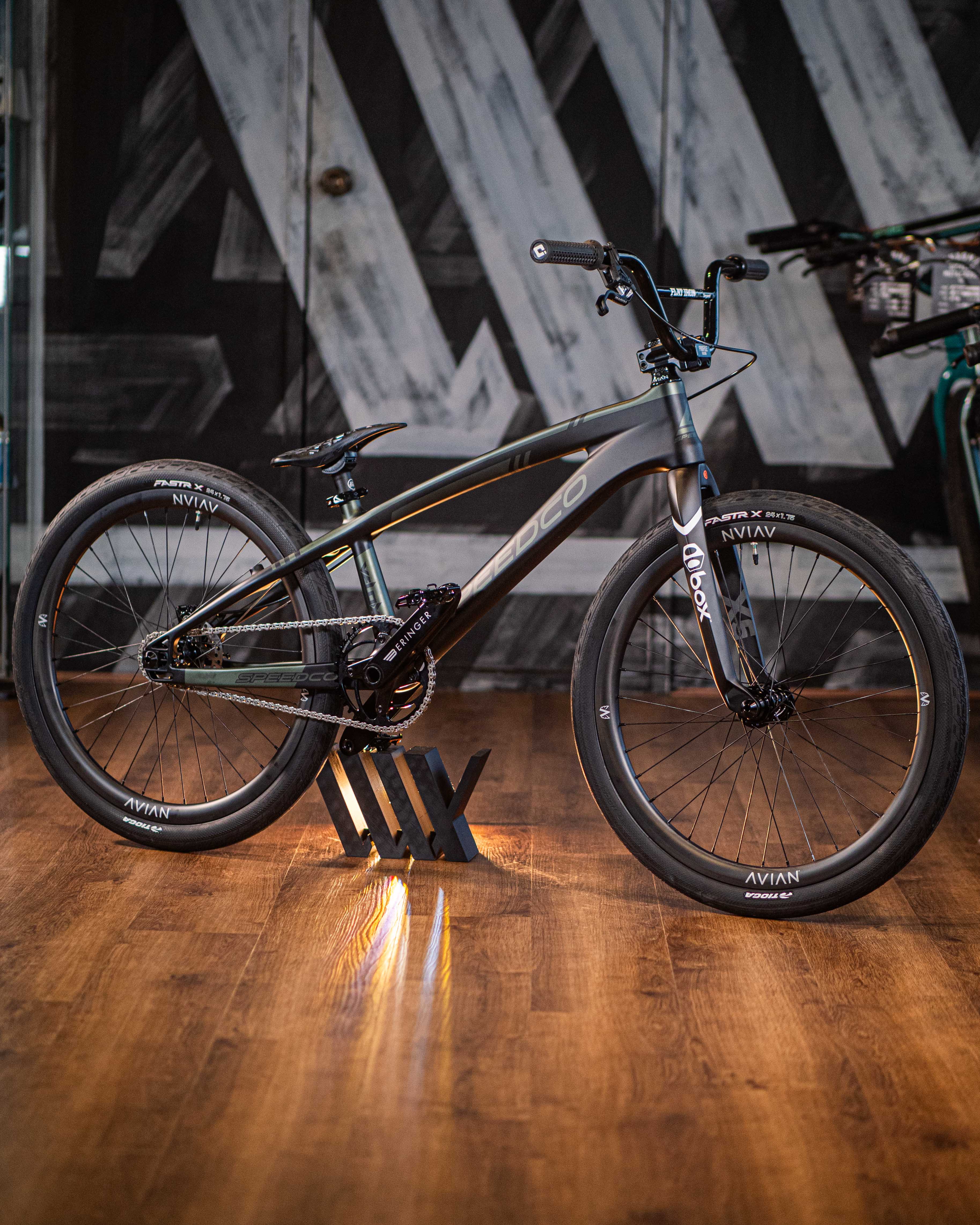 A sleek black Speedco Velox Pro XL Cruiser Custom Bike, featuring its distinctive angled top tube, is showcased indoors on a wooden floor against a backdrop of geometric wall patterns.