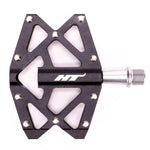 The HT AXO1A Nano Pedals are black with a metallic axle, featuring a geometric design and multiple grip pins. Ideal for young shredders, they include high-quality DU bushing for durability and have "HT" printed in white.