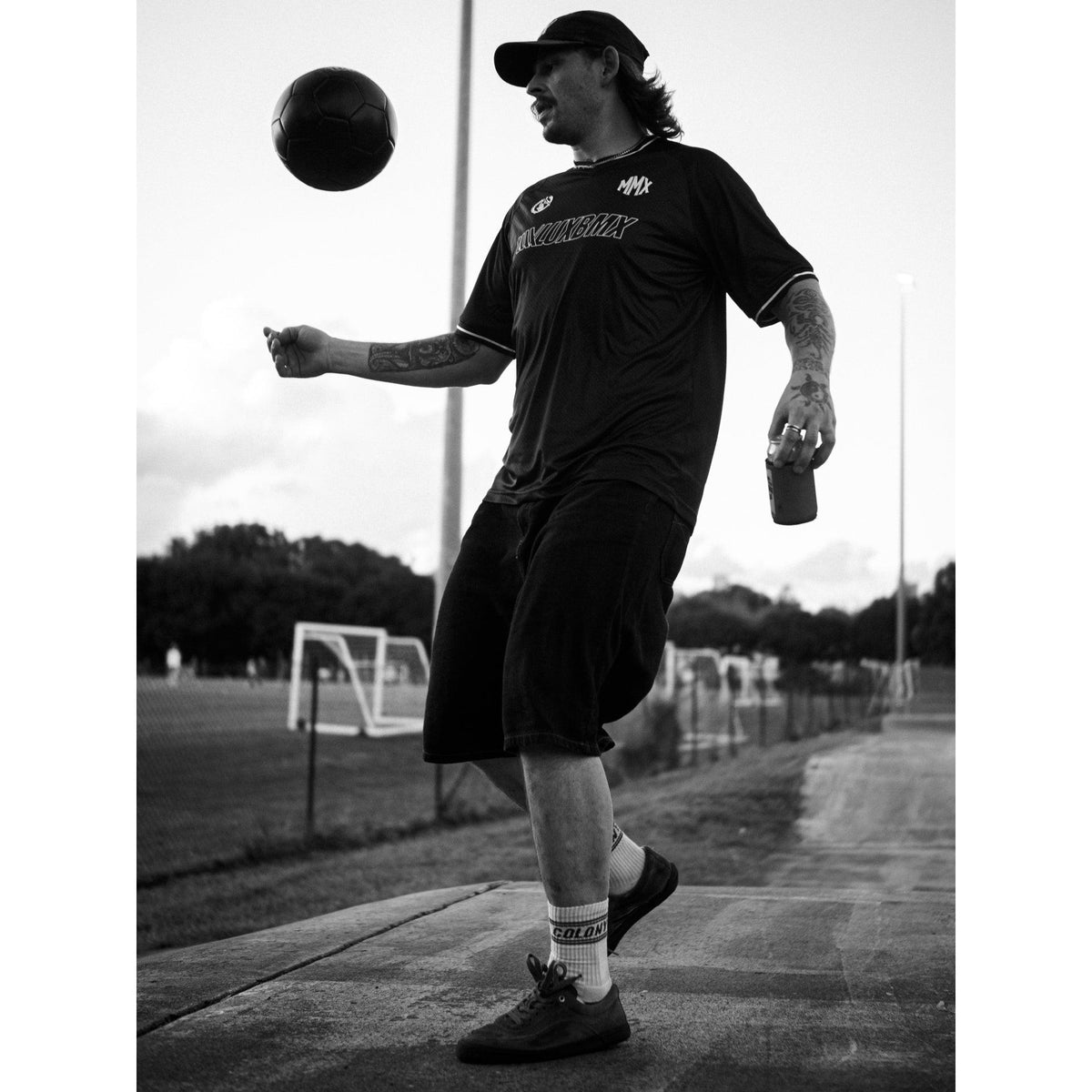 Wearing a LUXBMX MMX Soccer Jersey - Plain Back Print, a man juggles a soccer ball on one foot and holds a drink, enjoying the outdoors near soccer fields.