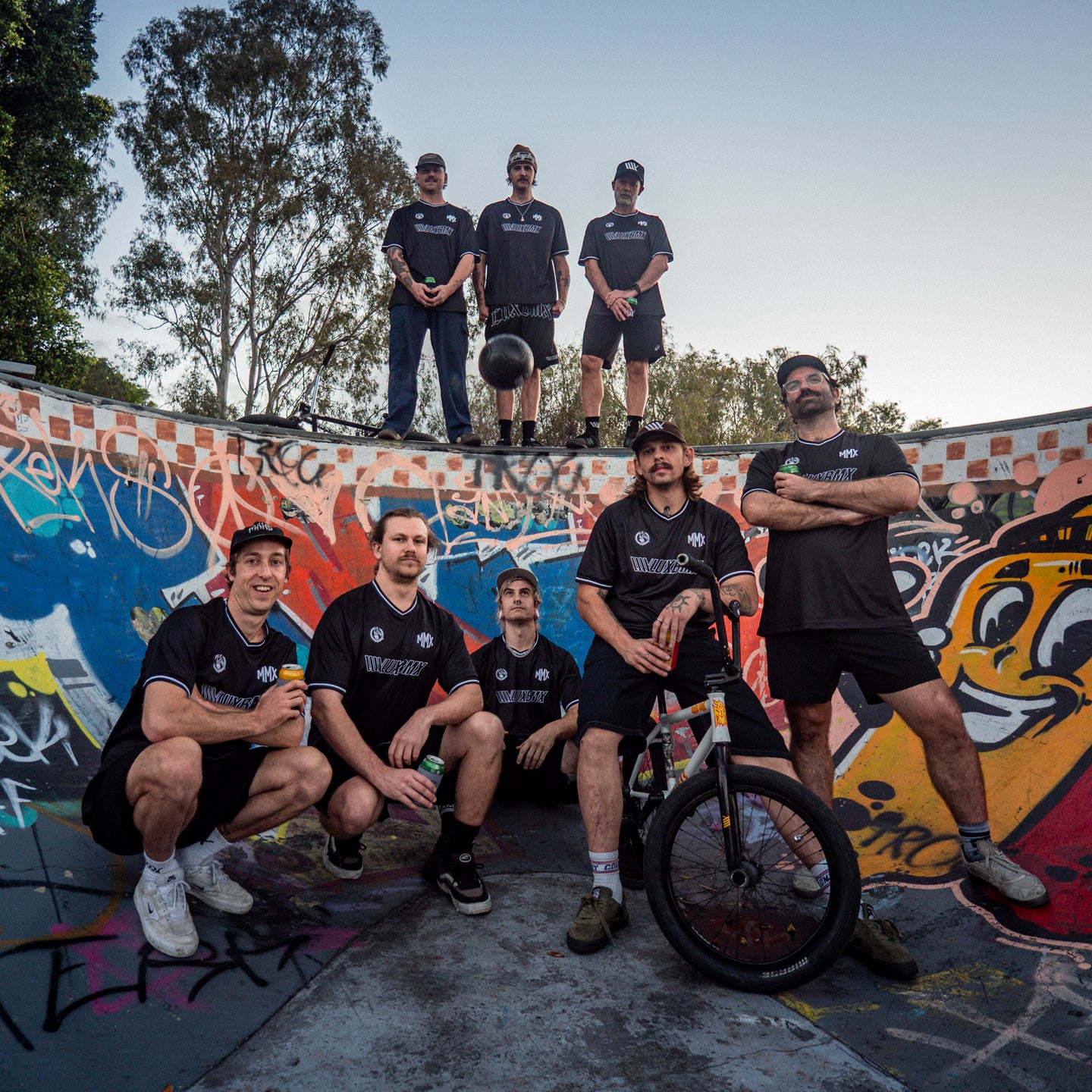 Eight men in matching dark athletic gear, including a standout LUXBMX MMX Soccer Jersey - Plain Back Print, pose in a colorful graffiti-covered skate bowl; some are standing while others sit casually, with one person holding a bike.
