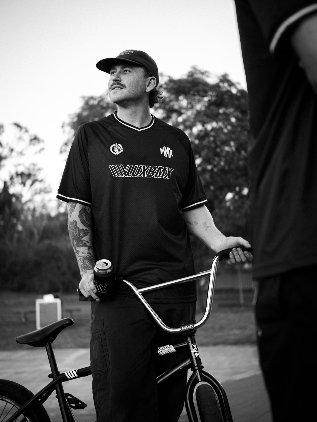 A man stands outdoors holding a BMX bike and a beverage can, wearing a LUXBMX MMX Soccer Jersey - Plain Back Print and a cap, with trees visible in the background.