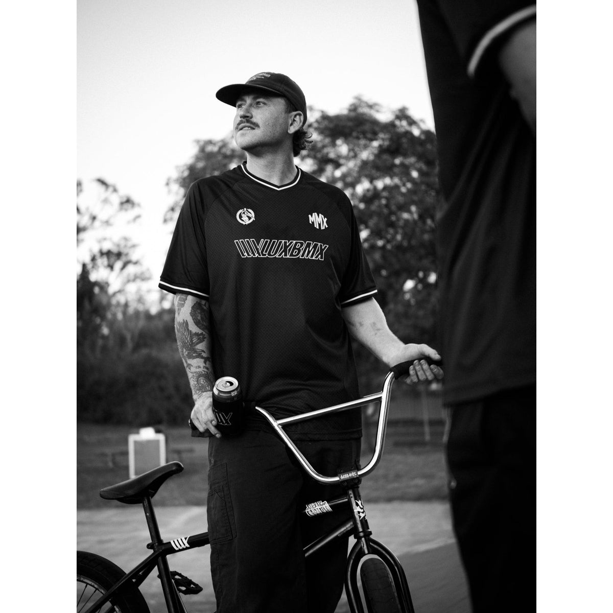 A man stands outdoors holding a BMX bike and a beverage can, wearing a LUXBMX MMX Soccer Jersey - Plain Back Print and a cap, with trees visible in the background.