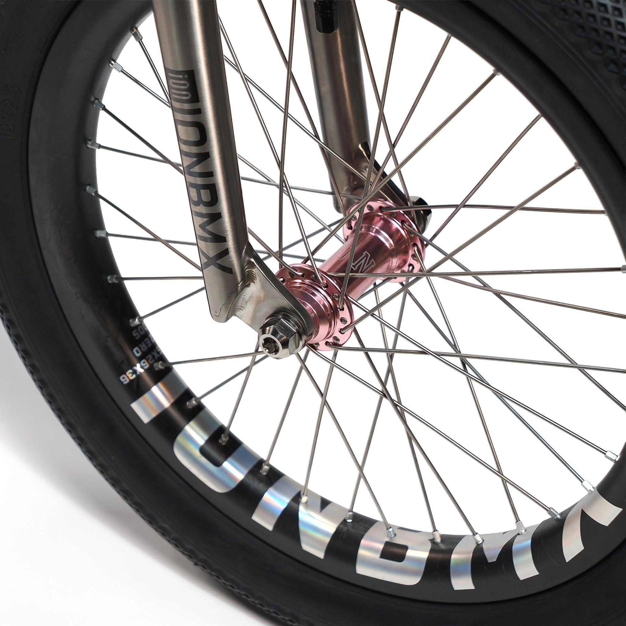 Close-up of the ION BMX Ti18 Custom Titanium 18 Inch Bike's front wheel, showcasing a pink hub, silver spokes, and a black tire with bold "DN BMX" branding—emphasizing its lightweight freestyle BMX build.