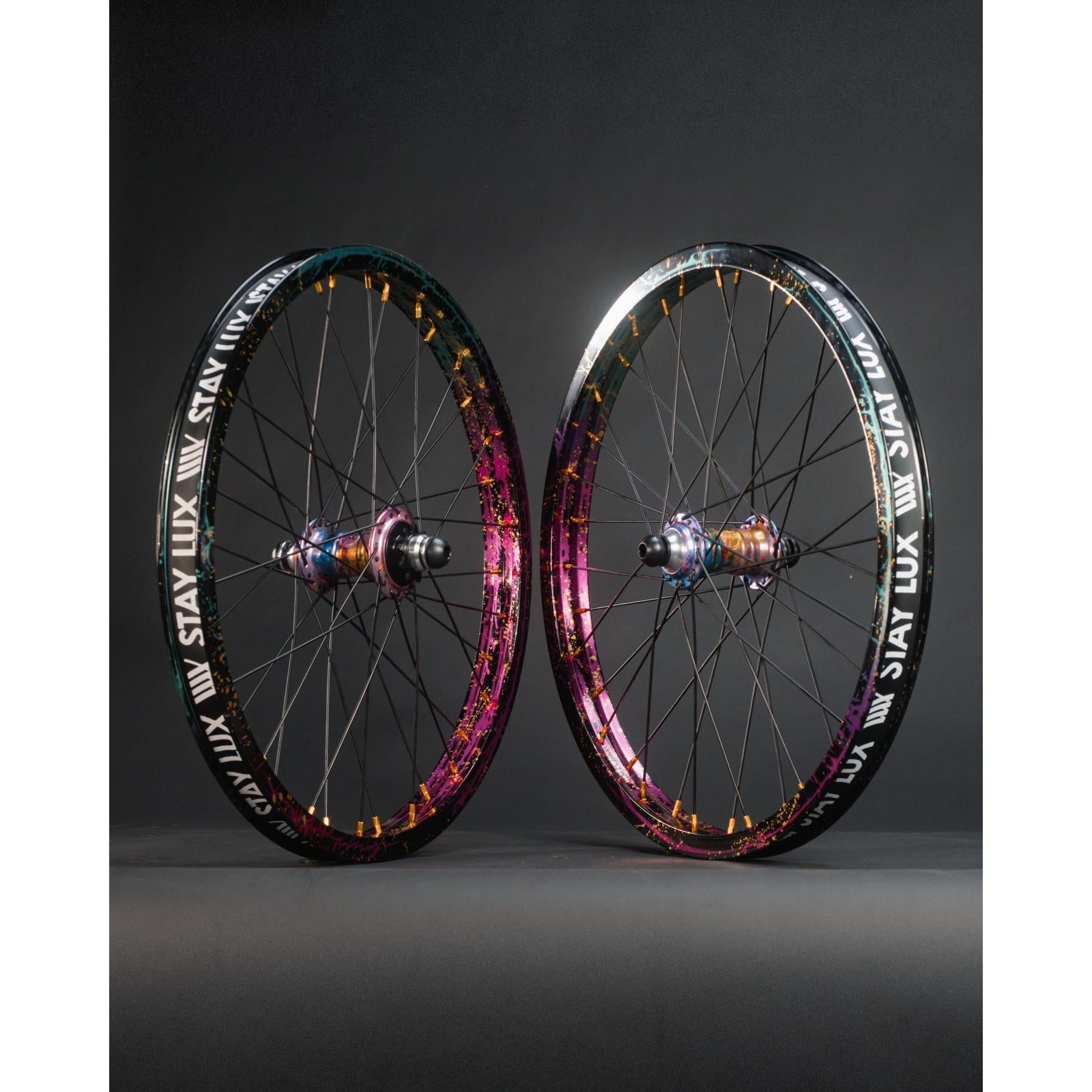 Two colorful bicycle wheels with black rims and metallic spokes, featuring BMX hubs, stand upright. The rims show “STAY LUX” in bold white text, highlighting the limited edition Profile x Odyssey Near Earth Objects Wheel Set.