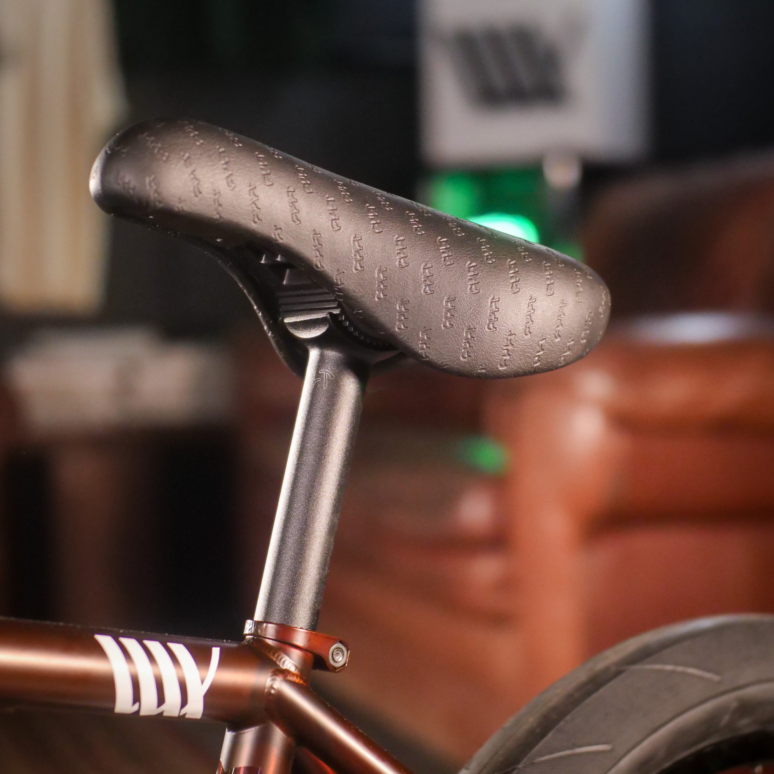 Close-up of a black bicycle seat and seat post on a Cult Dak X Eclat Custom 'Pat Freyne Signature' frame, set against a blurred indoor background.