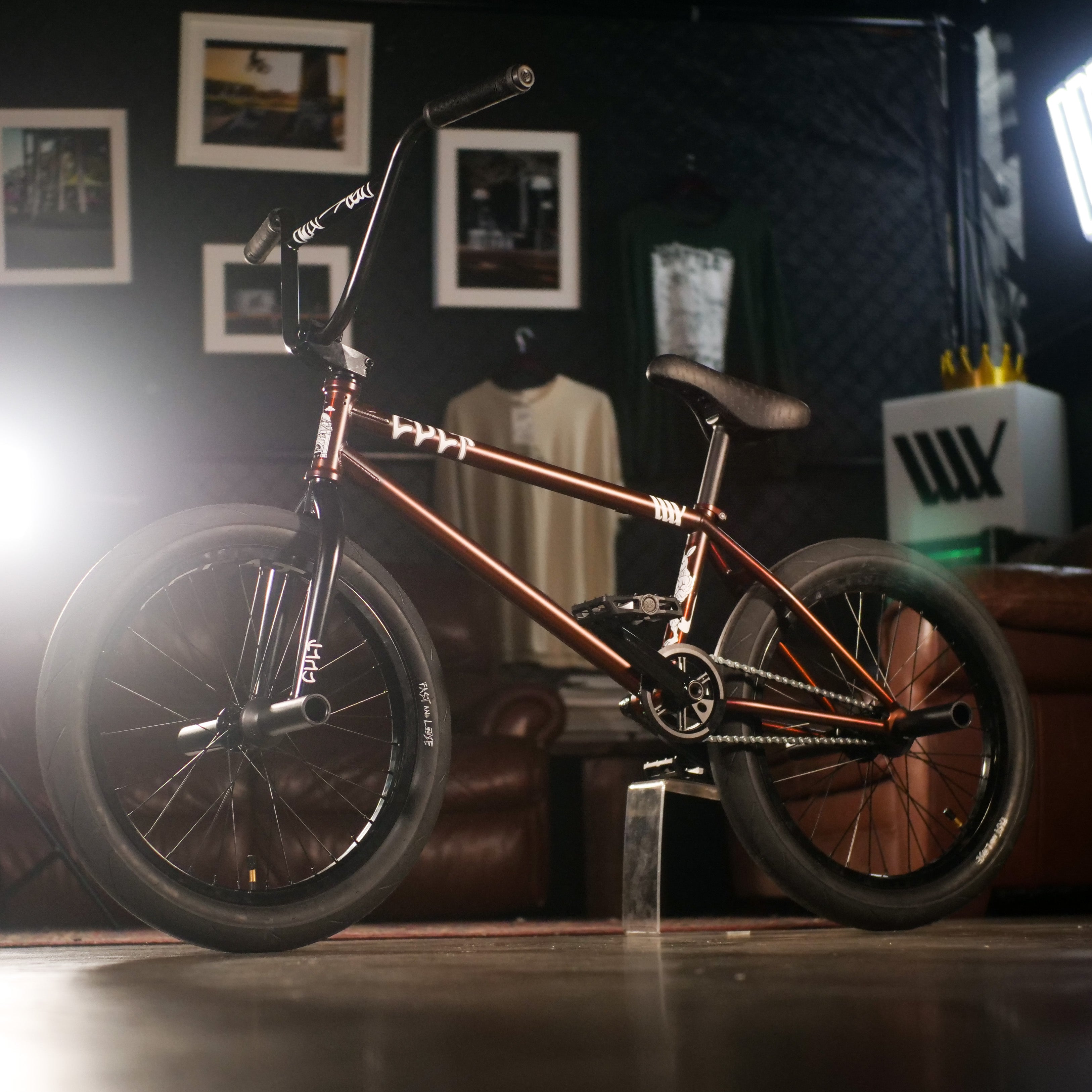 The Cult Dak X Eclat Custom 'Pat Freyne Signature' BMX bike, with a brown frame and black tires, is displayed indoors on a polished floor amid framed photos and furniture.