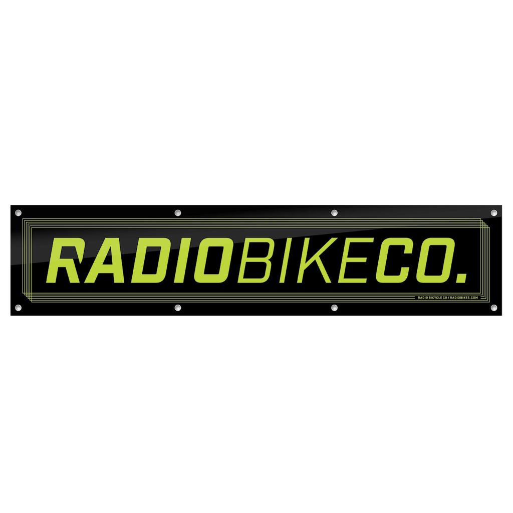 The Radio Contest Banner is a rectangular black PVC sign with yellow-green "RADIOBIKECO." text and edge mounting holes—ideal for outdoor use or displaying your favorite BMX brand.