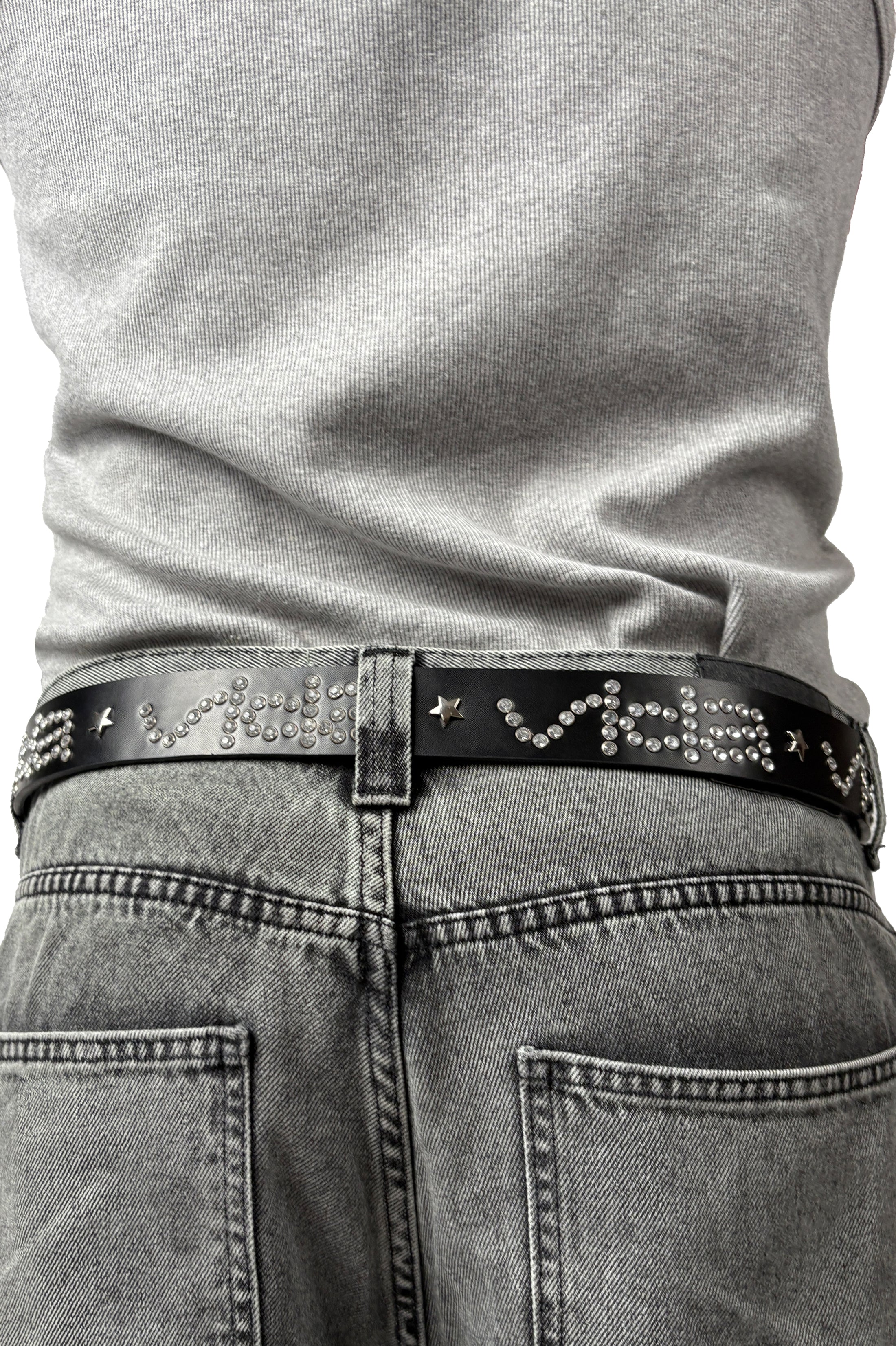 Person wearing a gray shirt tucked into faded gray jeans, styled with the V1da Studded Leather Belt featuring metallic text, star-shaped studs, and silver rivets for added edge.