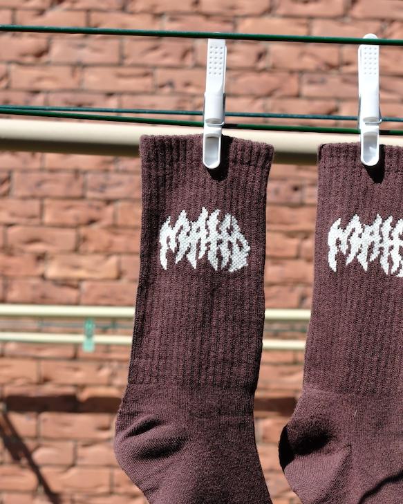 Mahd Metal Socks in a brown bamboo blend with white graphic text hang from a clothesline using plastic clips, set against a brick wall background. One size fits most.