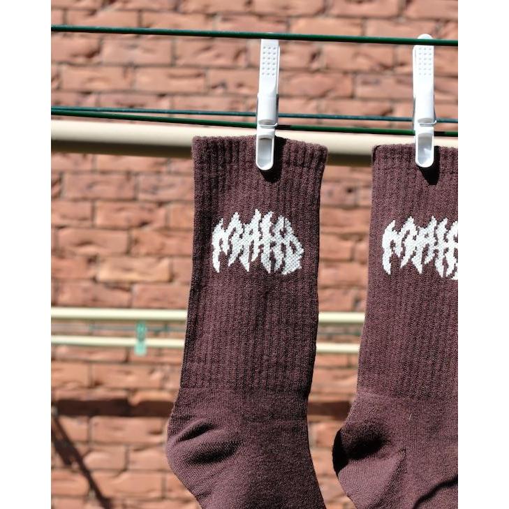 Mahd Metal Socks in a brown bamboo blend with white graphic text hang from a clothesline using plastic clips, set against a brick wall background. One size fits most.