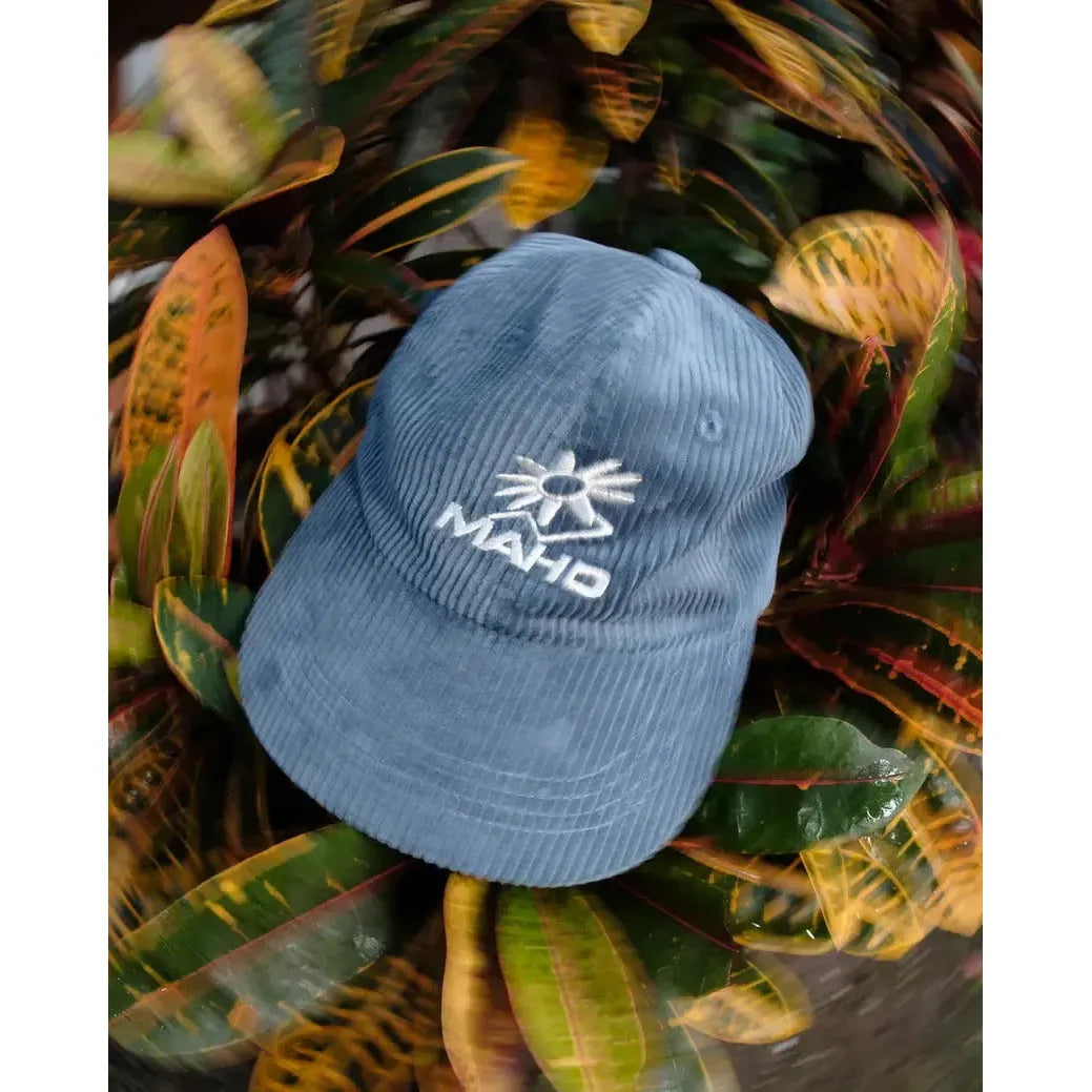 The Mahd Botanical Corduroy Hat, a blue cap featuring a white sun and “MAHD” logo, adds standout style as it sits atop colorful, variegated leaves.