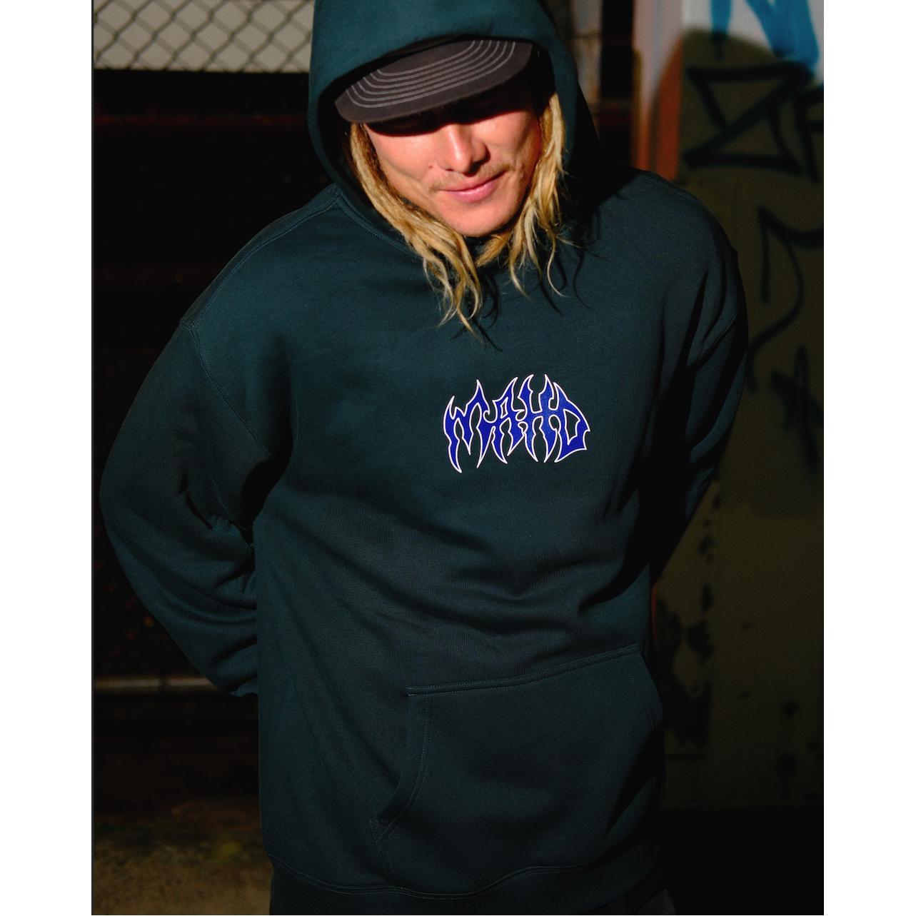 A person wearing the Mahd Metal Hoodie, featuring stylized "WORLD" text on the chest, stands outdoors at night by a chain-link fence with graffiti in the background.