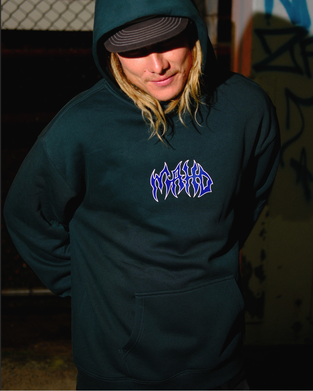 A person wearing the Mahd Metal Hoodie, featuring stylized "WORLD" text on the chest, stands outdoors at night by a chain-link fence with graffiti in the background.