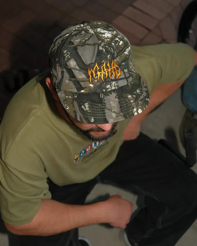 A person wearing the Mahd - Metal Hat, a green camouflage 5-panel cap with orange lettering, and an olive green shirt sits with their head slightly tilted down.