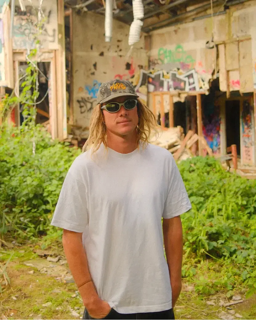 A person in a white t-shirt, sunglasses, and the Mahd - Metal Hat stands outdoors in front of an abandoned, graffiti-covered building surrounded by overgrown greenery.
