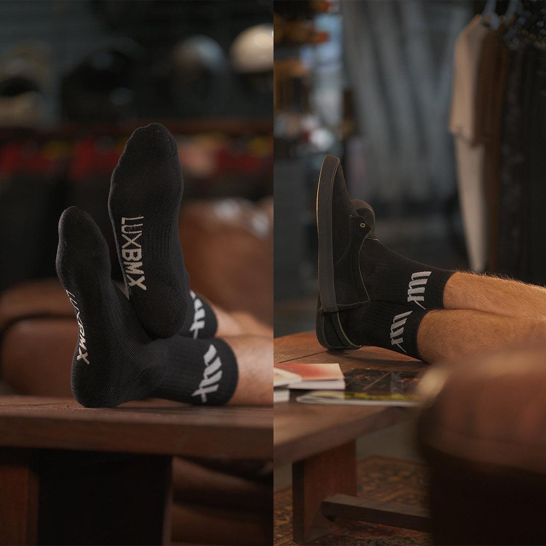 Two side-by-side images show a person relaxing indoors on a brown leather chair, feet on a table, wearing black LUXBMX Crew Socks with "LUX BMX" text.
