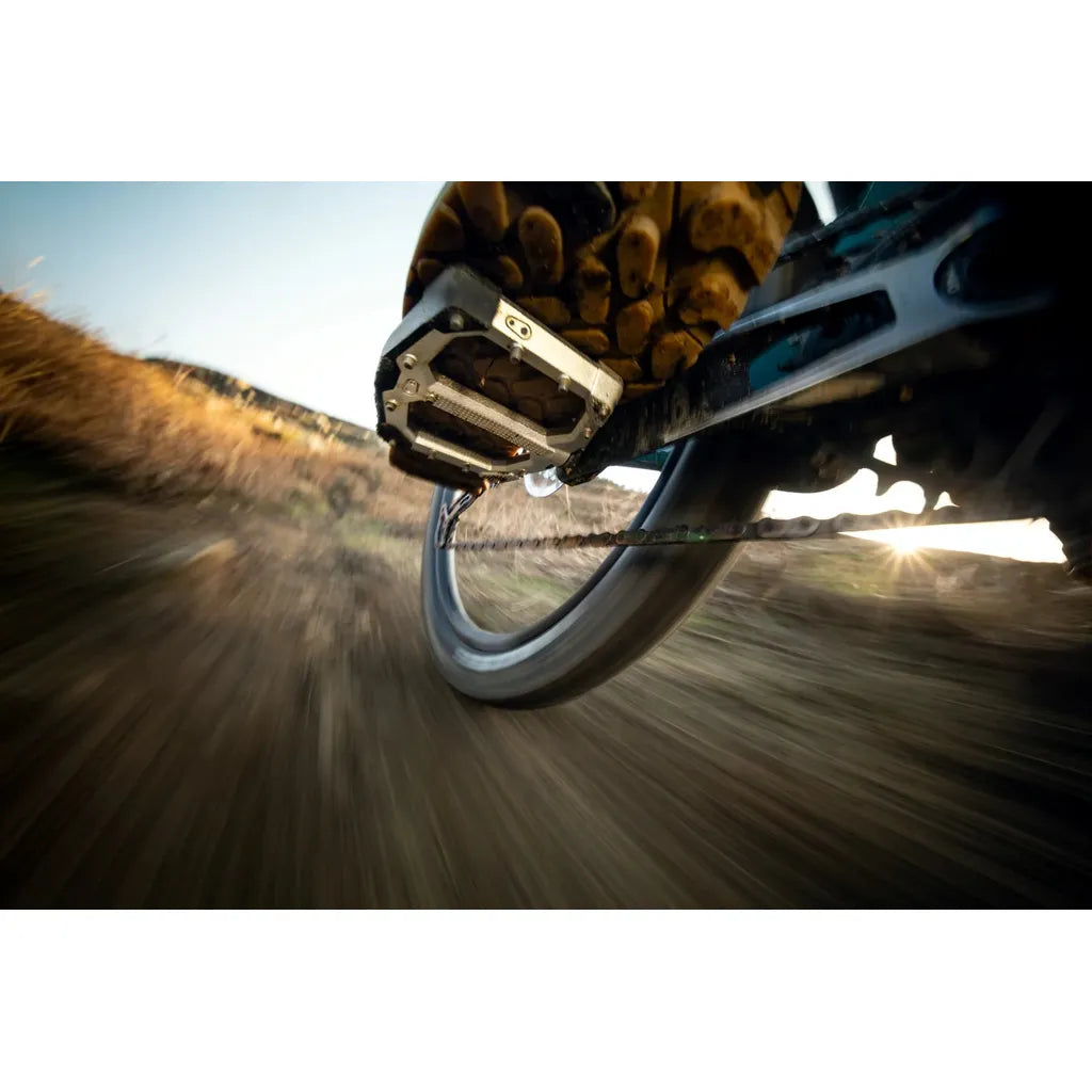 Close-up, low-angle view of a cyclist’s shoe on a large Crankbrothers Stamp Evo Pedals (Large) platform with adjustable pins while riding on a dirt trail.