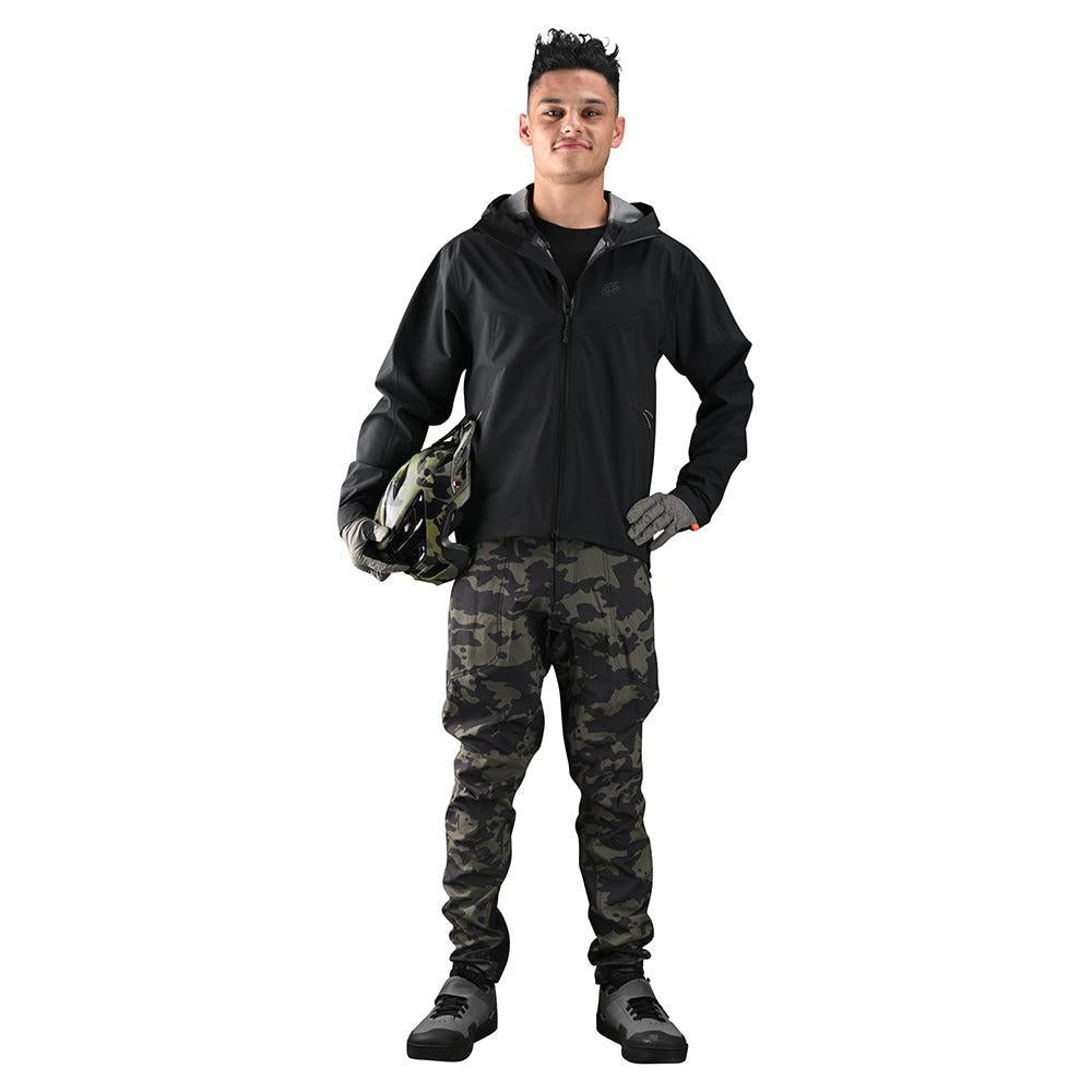 Man in a black TLD Descent Jacket and camouflage pants, holding a helmet and standing confidently against a white background.