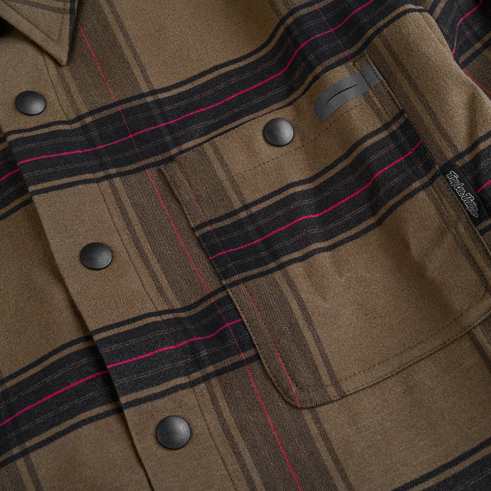 Close-up of the TLD 24.2 Grind Flannel Shirt in brown tech-plaid with black and pink stripes, featuring black snap buttons and a chest pocket. Experience quick-dry performance for all-day comfort.
