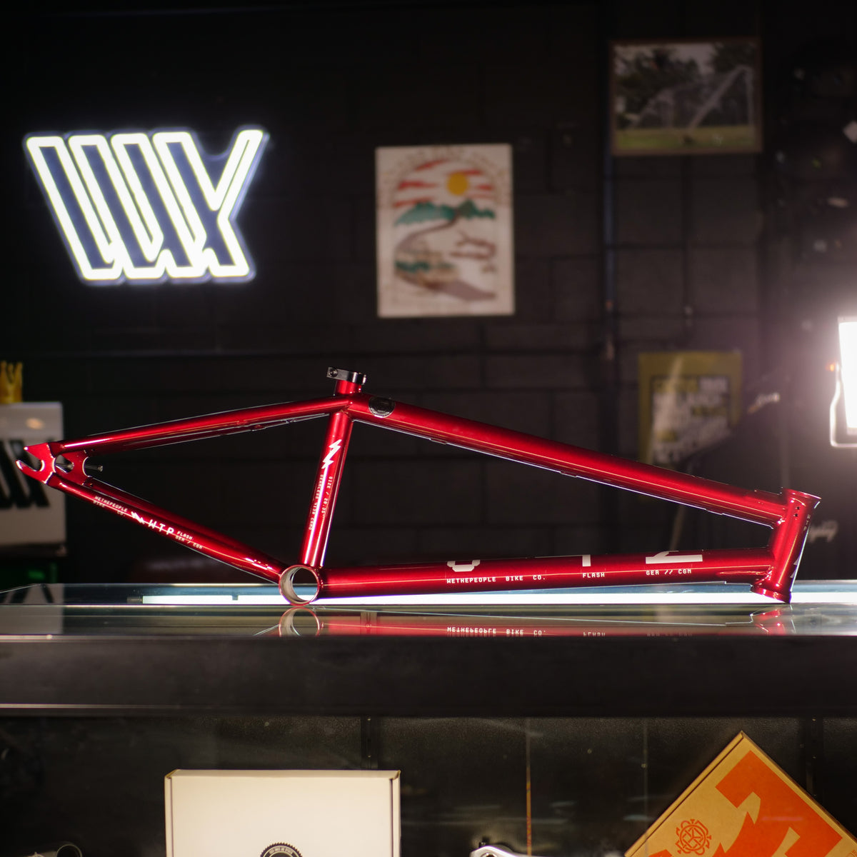 A Wethepeople Flash 20 Inch Frame in red, featuring a steep head tube angle, is displayed on a glass counter with various posters and a neon sign in the background.