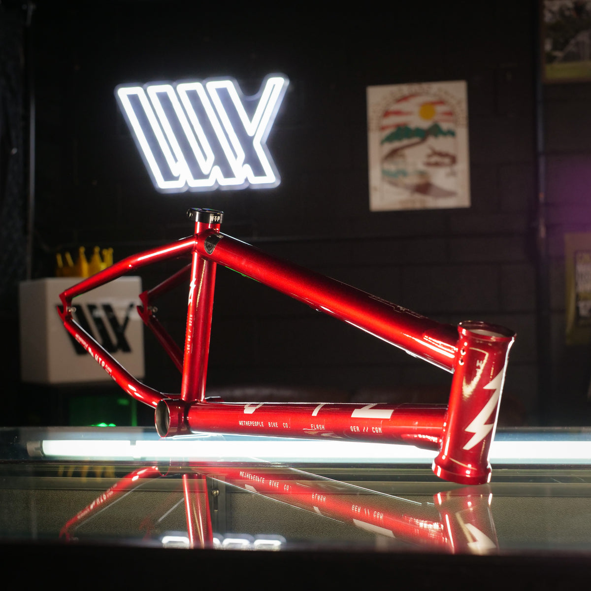 The Wethepeople Flash 20 Inch Frame, featuring a red finish and a white lightning bolt design, rests on a glass surface; its steep head tube suggests sharp handling, while a neon “WY” sign glows in the background.