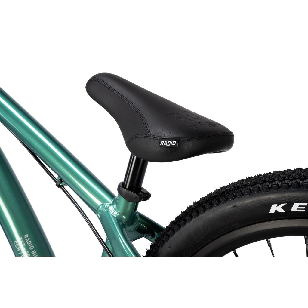 Close-up of a black saddle mounted on the Radio Griffin 24 Inch Dirt Jumper Bike’s metallic green frame, with part of a knobby tire and visible "KENDA" branding.