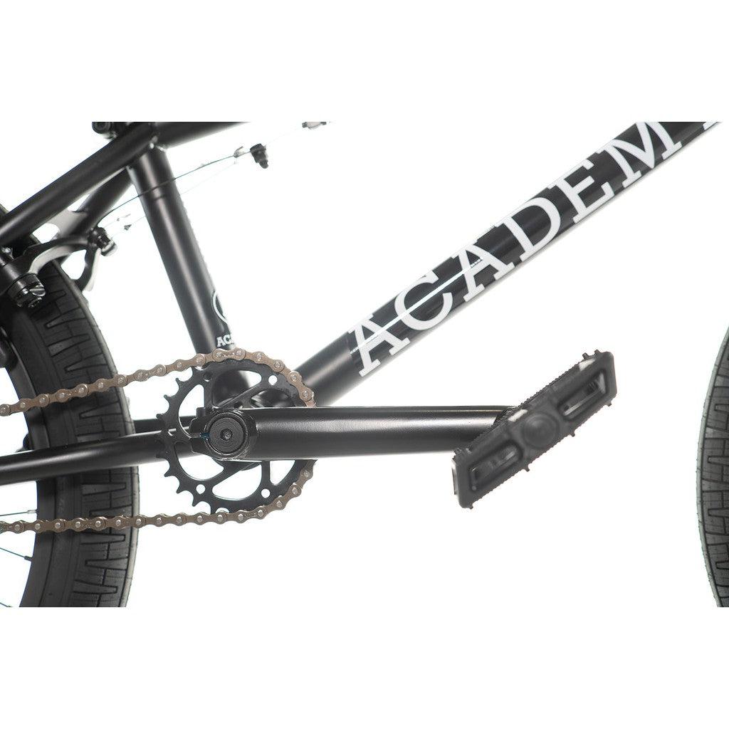 Close-up view of the chain, crankset, and pedals on the Academy Entrant 20 Inch Bike, showcasing the word "ACADEM" on the black freestyle BMX frame.