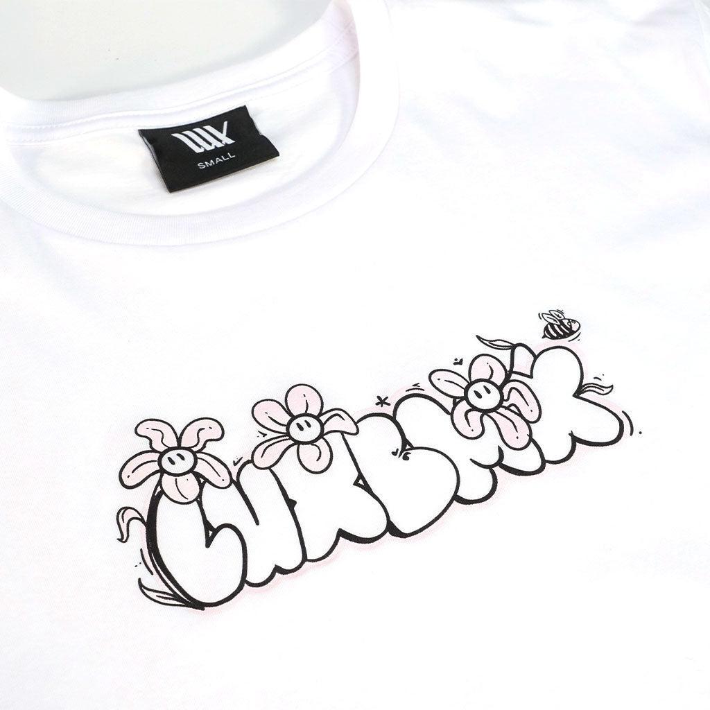 The LUXBMX Ali Crop Tee is a white cropped t-shirt with "CURBLICK" in bubble letters, cartoon flowers, and a small bee above the text. Made from a breathable cotton blend for all-day comfort.