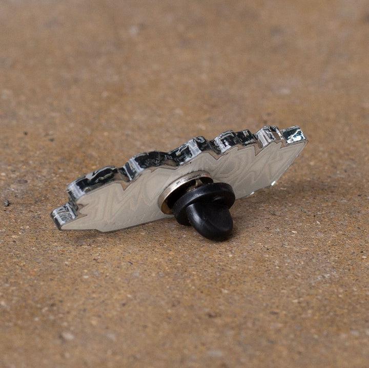 The Backbone Always Building Pin, featuring a black rubber backing, elegantly rests on a textured brown surface, showcasing top-tier softgoods design.