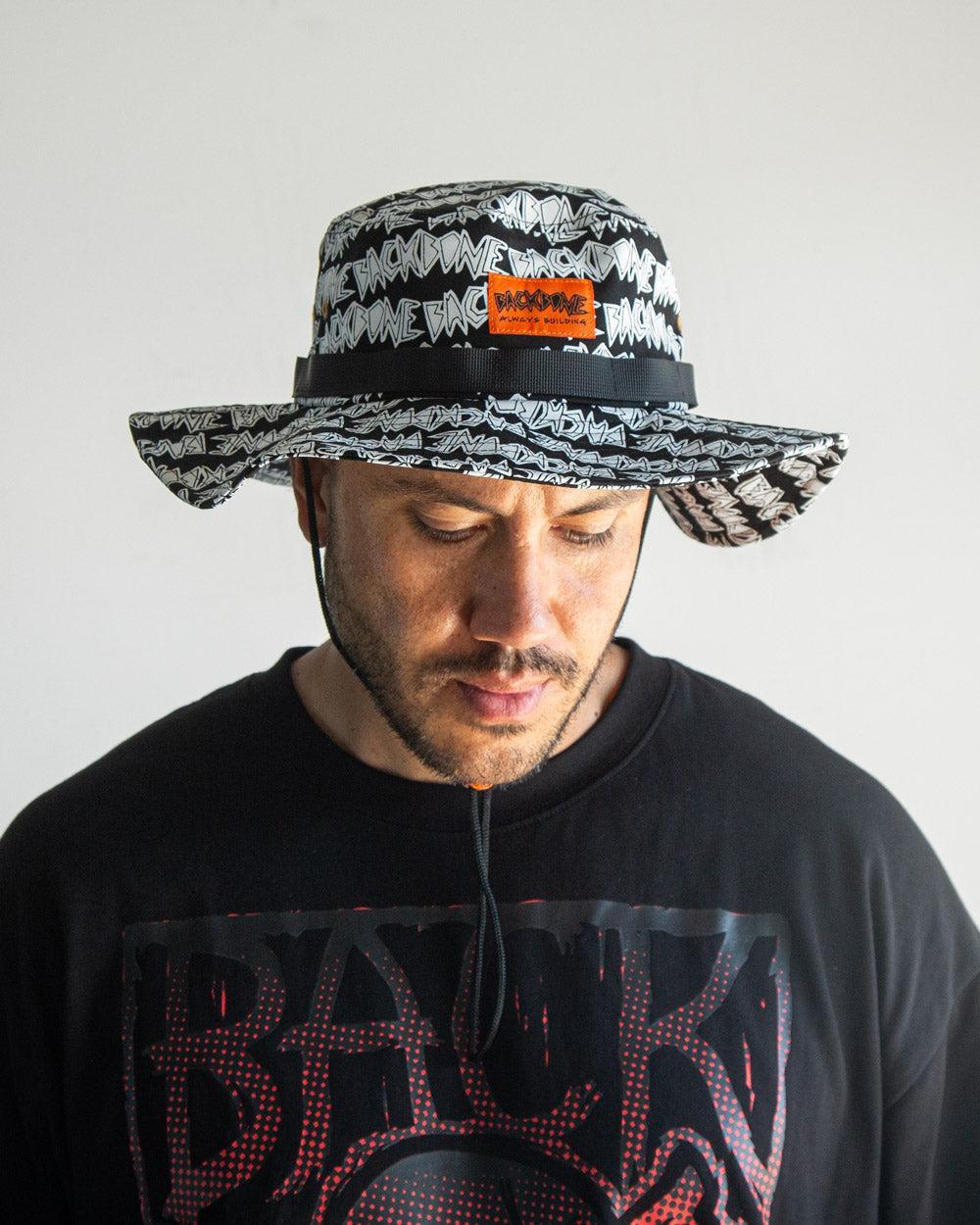 Man wearing the Backbone AO Boonie, a wide-brimmed black and white bucket hat with repeating text and an orange patch, paired with a black graphic tee against a plain background.