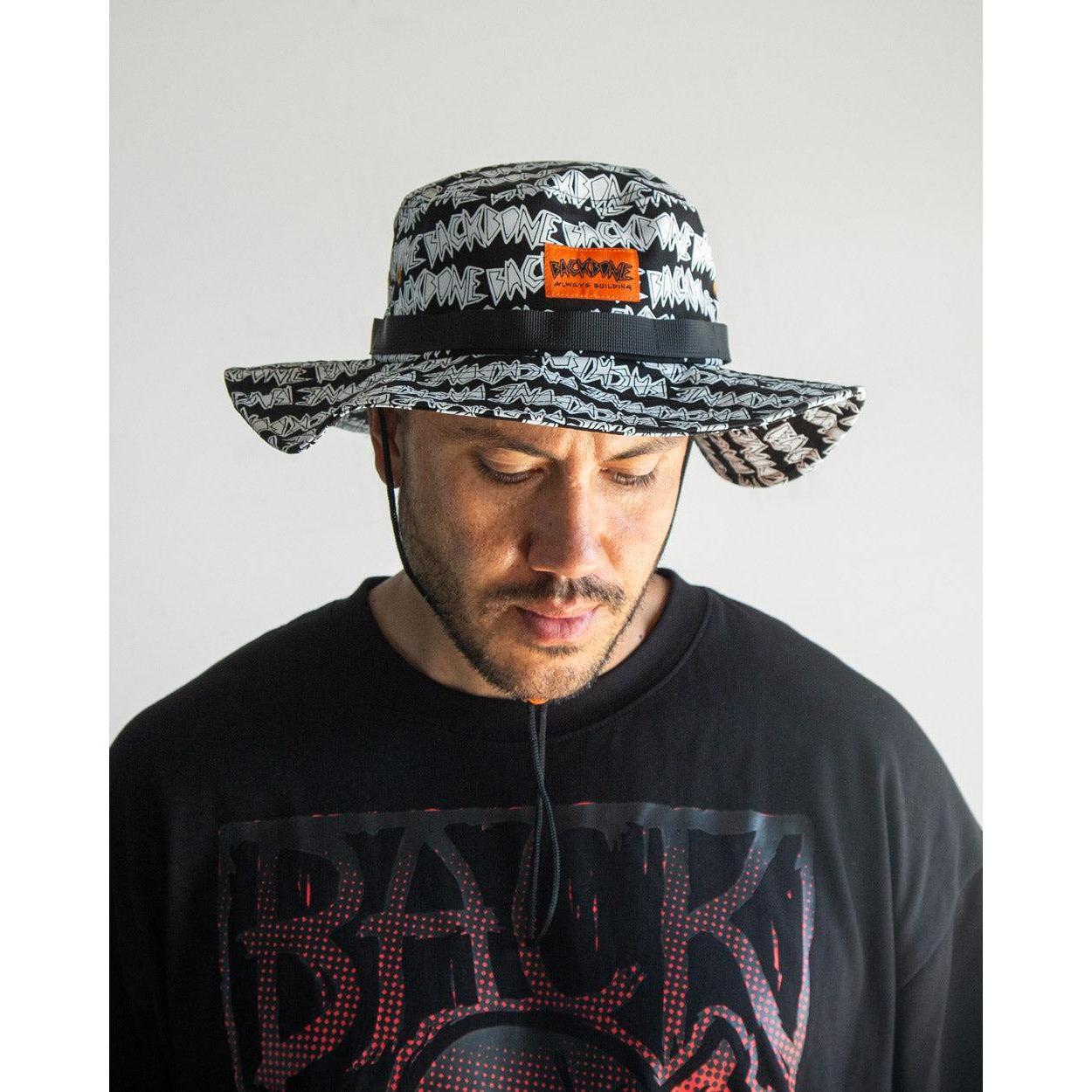 Man wearing the Backbone AO Boonie, a wide-brimmed black and white bucket hat with repeating text and an orange patch, paired with a black graphic tee against a plain background.