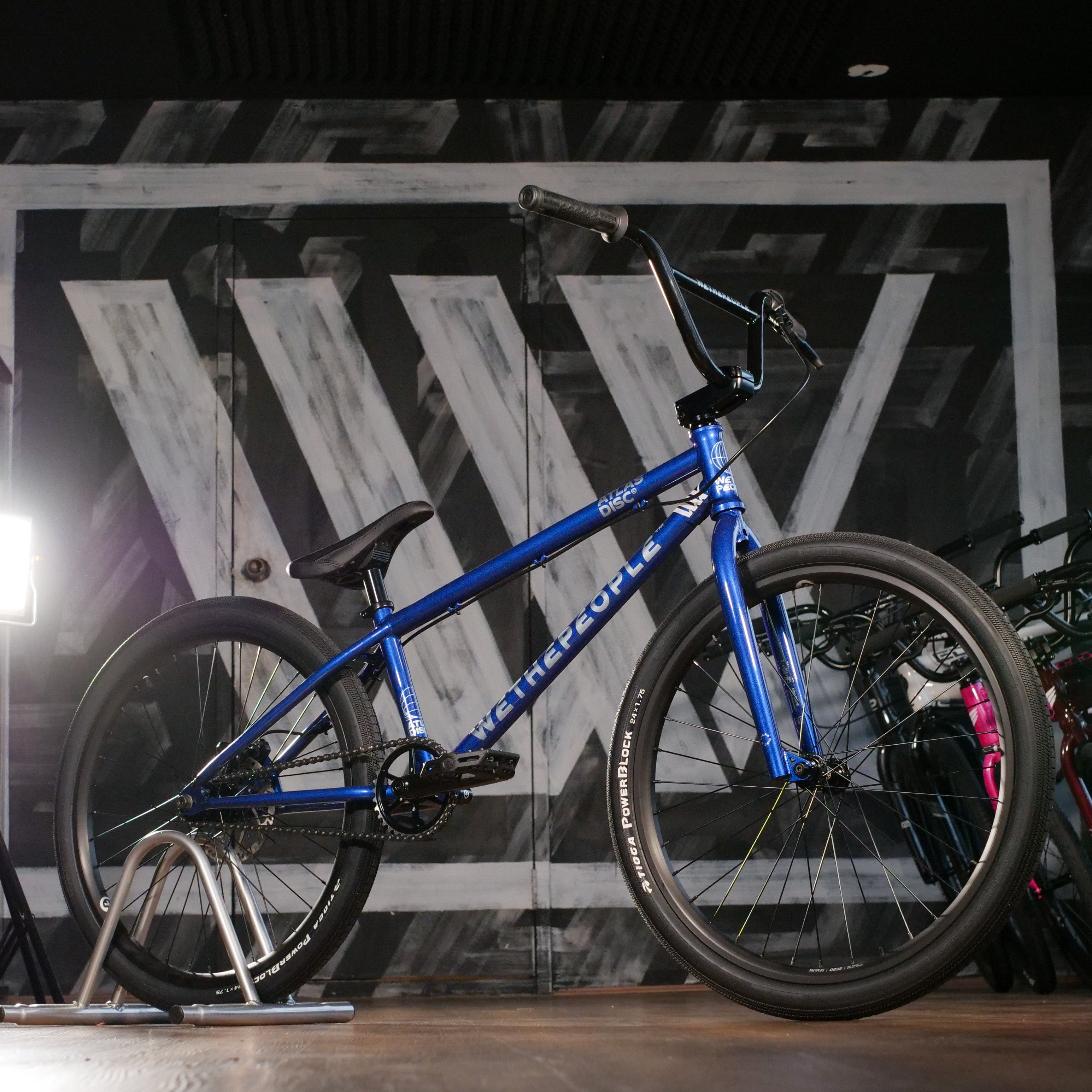 The Wethepeople Atlas Disc 24 Inch Bike, featuring a blue frame with race-inspired geometry, black tires, and handlebars, is displayed indoors on a stand with a geometric black and white mural in the background.
